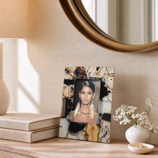 A rectangular picture frame with an agate geode pattern in black, brown, and cream, holding a portrait of a woman with long dark hair and gold earrings. The frame is styled on a wooden table next to a lamp, books, and a vase of white flowers.