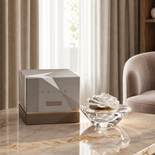 A luxury home fragrance product shown with its metallic silver and gold box on a polished marble table, next to a clear glass diffuser with a white rose-shaped cap in a softly lit living room.