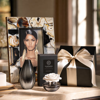 A luxury gift set arranged on a marble surface in a warm, sunlit interior. The set includes a black and white rose diffuser, a black metallic vase, a black gift box tied with a cream ribbon, and a black rectangular box labeled EKLUMARIA.