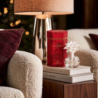 A red rectangular gift box with gold detailing and a small clear glass diffuser on stacked books beside a lamp in a cozy indoor setting.