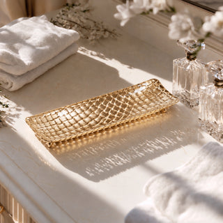A rectangular gold tray with a textured, woven basket-like pattern, styled on a marble bathroom counter alongside white towels and clear glass perfume bottles.