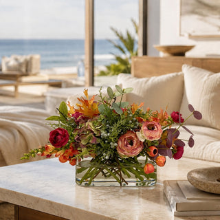 A vibrant floral arrangement in a clear glass rectangular vase, set on a marble coffee table in a bright, modern living room with a beach view in the background.