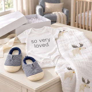 Lifestyle photo of the baby boy essentials gift box showing the white gift box, striped blue and white faux leather booties, so very loved onesie ad cotton muslin blanket/swaddle styled in a contemporary nursery.