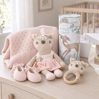 Biggest Little Baskets photo of an heirloom A baby gift set arranged on a light wood dresser in a softly lit nursery. The set includes a plush deer doll in a pink dress, a pink knit blanket, a pair of pink crocheted booties, a wooden teething ring with a deer face, and a cylindrical gift box printed with 'Forest Fawn' and deer illustrations.
