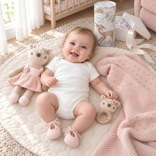 Styled photo of a baby girl with the Heirloom Baby Girl Gift box. Forest Fawn toy, forest dawn rattle with wood teether, pink heirloom knit blanket, pink crocheted booties and a white keepsake gift box  from Biggest Little Baskets