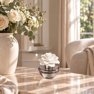 A small rose-shaped diffuser or fragrance atomizer in a clear round base sits on a marble table in a softly lit living room, with a large vase of white roses and greenery beside it.
