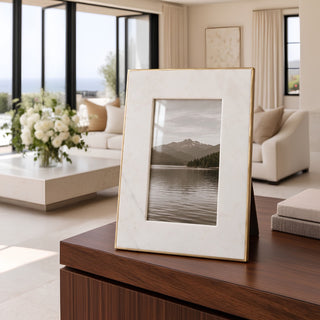 A vertical picture frame with a white marble-look border and a thin gold metal frame, standing on a dark wood table in a modern, sunlit living room. The center is blanked out for privacy.
