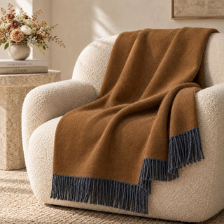 Tapenade bronze-brown throw with charcoal fringe draped over a cream armchair beside a stone table and flowers.