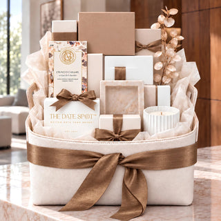 A large curated faux leather gift basket arrangement on a marble surface, featuring multiple gift boxes, tissue paper, a candle, and decorative branches, with visible brands including The Date Spot and Garcia Nevett. For corporate gifting and luxury personal gifting or celebrations, bereavement, congratulations and housewarming gifts from Biggest Little Basets