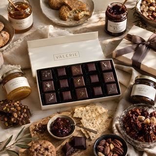 A Styled photo of Biggest Little Baskets $350 Gifting Tier custom curated gift box gourmet gift box of chocolates and spreads from Valerie, displayed in a beautifully styled flat-lay arrangement with cookies, jars of jam, crackers, and nuts on a neutral fabric background.
