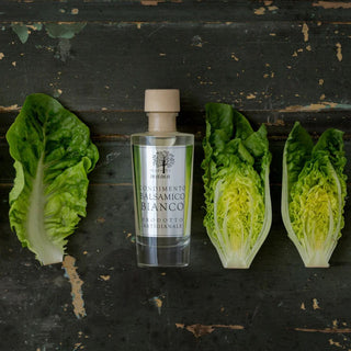 Clear bottle of white balsamic Bianco styled with romaine leaves on a dark wood surface.