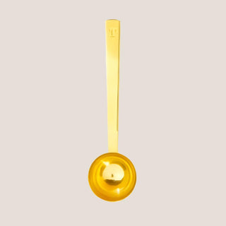 Brushed gold single-serving tea scoop shown alone against a warm neutral background.