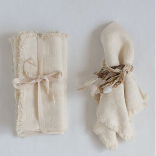 Two styling views of soft cream linen-blend napkins with fringe, one tied as a set and one gathered with a botanical sprig.
