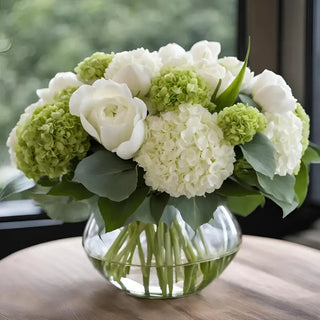 Green and white real-touch hydrangea and rose arrangement displayed in a round clear glass vase beside a window.