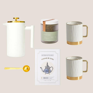 Flat lay of the Luxe Tea Indulgence collection with white-and-gold French press, two mugs, Green Gold tea, cookies, and gold scoop.