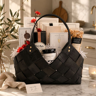 The Marlowe black woven gourmet gift basket with artisan foods and red botanical accents styled on a marble kitchen counter.