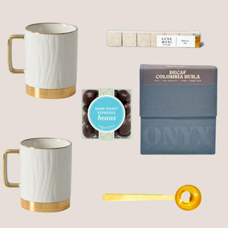 Flat lay of Petite Java Joy with two white-and-gold mugs, Decaf Colombia Huila coffee, espresso beans, vanilla cubes, and gold scoop.