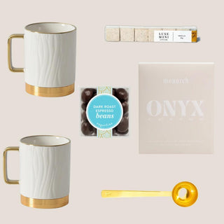 Flat lay of Petite Java Joy with two white-and-gold mugs, Monarch whole-bean coffee, espresso beans, vanilla sugar cubes, and gold scoop.