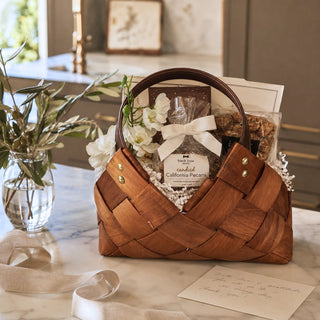 Petite woven gourmet gift basket filled with sweet and savory treats and a seasonal botanical accent on a marble surface.