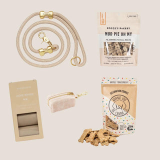 Flax rope leash, canvas waste-bag dispenser, compostable bags and two gourmet dog treats arranged on a warm neutral background.