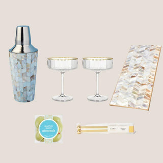 Flat lay of the Shaken, Not Stirred collection with shell mosaic shaker and tray, two gold-rimmed coupes, olive almonds, and gold picks.