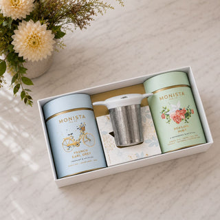 Open Tea Lovers’ Bliss gift box with French Earl Grey and Persian Mint tea canisters, a metal stick infuser, and cream flowers on a marble surface.
