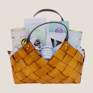 Front view of the medium woven Tea Time Gift Basket with two tea canisters, artisan treats, a gold scoop, and dark handles.