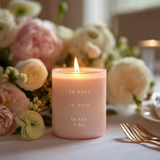 Lit blush To Have, To Hold wedding candle with gold lettering styled among soft wedding flowers and tableware.