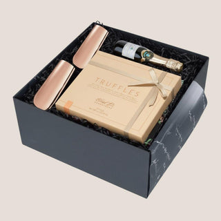 Angled view of the open black Truffles & Bubbles box with dark fill, copper flutes, truffles and bottle candle.