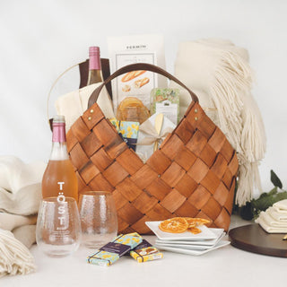Ultimate Home Basket displayed with its woven basket, cream throw, alcohol-free sparkling tea, hammered glasses, white plates, chocolates, and serving pieces.