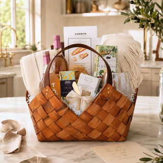 Large woven Ultimate Home Gift Basket with throw, serving pieces, sparkling tea, glasses, plates, and gourmet treats on a marble kitchen counter.