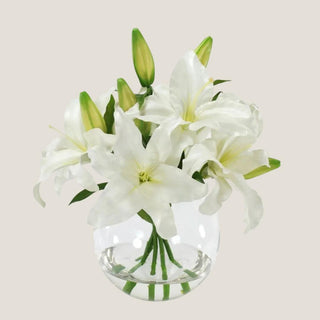 Lifelike white Casablanca lilies and green buds arranged in a rounded clear glass vase with realistic faux water.