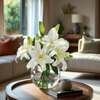 White Casablanca lily faux floral arrangement in a round clear glass vase styled on a sunlit living-room coffee table.
