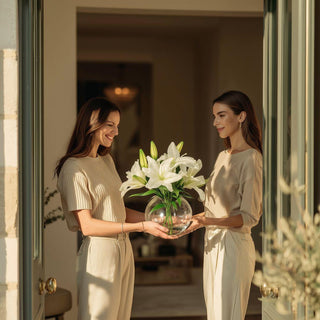 Two women exchanging a white Casablanca lily faux floral arrangement in a clear glass vase at a home's doorway.