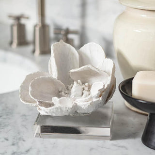 White magnolia resin sculpture on a clear crystal base styled on a marble bathroom counter beside soap and ceramic jars.