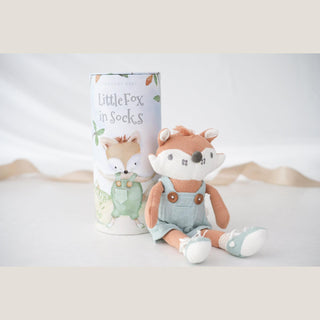 Rust and muted green knit fox toy seated beside its illustrated Little Fox in Socks cylinder on a softly lit white surface.