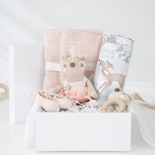 Open white Heirloom Baby Girl gift box with pink cotton knit blanket, ballerina booties, fawn wooden ring rattle, knit fawn toy, and illustrated story cylinder.