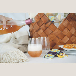 Alcohol-free pink sparkling tea being poured into a hammered stemless glass beside a second glass, chocolates, plates, and woven basket.