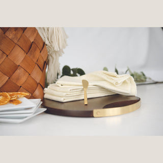 Folded cream napkins and a matte gold cheese spreader arranged on a dark round mango wood serving board.