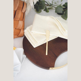 Dark mango wood serving board with gold-tone handle, folded cream napkins, and matte gold spreader beside the woven basket.
